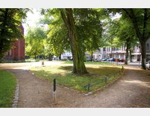Ein kleiner Park mit Kieswegen, einer Rasenfläche und großen, schattenspendenden Bäumen. Im Hintergrund befinden sich mehrstöckige Wohnhäuser und geparkte Autos; links ist die Fassade eines roten Backsteingebäudes zu sehen.