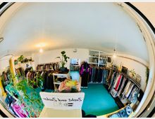 Kleiner Second-Hand-Laden mit bunten Kleidungsgestellen entlang der Wände, türkisfarbenem Boden und Zimmerpflanzen. Das Bild ist durch einen gewölbten Spiegel aufgenommen, der den ganzen Raum und die Einrichtung reflektiert.