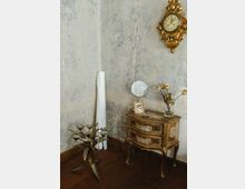 Antike Zimmerecke mit einer vergoldeten Wanduhr, einer verzierten Kommode im Barockstil, darauf ein kleiner Spiegel und ein Bilderrahmen, sowie getrockneten Blumen in einer Glasvase. Daneben steht ein florales Metallobjekt mit kerzenförmigen Lampen vor einer abgenutzten und rissigen Wand.