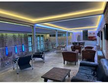 Industrieller Innenraum mit einer Mischung aus modernen und Vintage-Möbeln, darunter Ledersessel, ein Sofa und ein Couchtisch auf Betonboden. Wände aus unverputztem Ziegelstein, Glasgeländer und eine indirekte Deckenbeleuchtung mit gelben und blauen LED-Streifen prägen das Design.