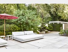 Modern gestalteter Gartenbereich mit großzügigen weißen Polsterliegen auf hellen Steinplatten. Im Hintergrund befinden sich eine Hecke, ein kleiner Pavilion und dicht bepflanzte Bäume, sowie ein rosafarbener Sonnenschirm und ein japanisch anmutender Baum.