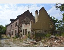 Altes, teilweise verfallenes Gebäudeensemble aus Backstein und Putz mit kaputten Fenstern, eingestürzten Wänden und Schutthaufen im Vordergrund; umgeben von vereinzelter Vegetation und einem Kopfsteinpflasterweg.