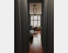 Ein schmaler Durchgang mit grauen Vorhängen führt in einen Raum mit Holzboden, einem großen Fenster mit weißem Rahmen und einem Kronleuchter an der Decke. Im Raum stehen ein Sofa, ein kleiner Tisch und ein Heizkörper unter dem Fenster.