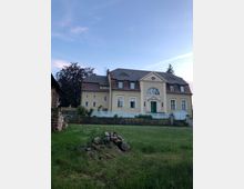 Herrenhaus im klassischen Stil mit hellbeiger Fassade, symmetrischem Aufbau und einem grünen Eingangstor, gelegen auf einem leicht erhöhten Fundament mit umliegendem Garten. Im Vordergrund befindet sich eine Grasfläche mit einem Steinhaufen, im Hintergrund Bäume und ein blauer Himmel.