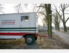 Ein Parkplatz mit einem Anhänger im Vordergrund, auf dem "Traberennen in Mariendorf" steht. Im Hintergrund sind Bäume, weitere parkende Anhänger und ein Weg zu sehen.