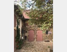 Ein Innenhof mit Kopfsteinpflaster, umgeben von einem historischen Gebäude aus Backstein mit rotem Ziegeldach. Zwei rote Holztüren sind in der Backsteinwand, daneben wachsen Pflanzen; ein Baum dominiert den Vordergrund.