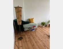 Wohnzimmer mit hellem Holzboden, einem grünen Polstersessel mit gelbem Kissen, einer stehenden Vintage-Lampe und einem kleinen Glastisch mit Büchern. Im Hintergrund stehen mehrere Zimmerpflanzen.