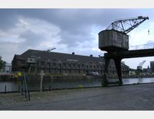 Industrielle Hafenlandschaft mit einem großen, alten Kranturm auf der rechten Seite und mehreren Backsteingebäuden im Hintergrund entlang eines Kanals. Im Vordergrund sind Gleise und gepflasterte Wege zu sehen, die zu Lagerbereichen führen.