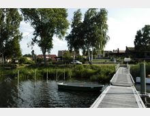 Ein Steg aus Holz führt über ein Ufergewässer mit einem kleinen Ruderboot und endet an einer grünen Wiese mit einzelnen dichten Bäumen. Im Hintergrund sind mehrere Wohnhäuser mit roten und braunen Dächern sowie ein geparktes Auto zu erkennen.