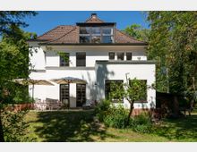 Rückansicht eines zweigeschossigen weißen Wohnhauses mit braunem Ziegeldach und großer Gaube mit Fensterfront, umgeben von Bäumen. Im Garten davor liegen eine Terrasse mit Sonnenschirmen und Sitzmöbeln sowie Rasen, Sträucher und ein kleiner Baum.