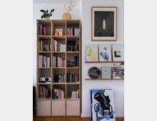 Ein modern eingerichtetes Wohnzimmer mit einem hohen Regalsystem, das Bücher, Dekorationsgegenstände und einen kleinen Projektor enthält. Daneben befinden sich schmale Wandregale mit Kunstbüchern und Bildern sowie weitere auf dem Boden gelehnte abstrakte Gemälde; an der weißen Wand hängt gerahmte Kunst.