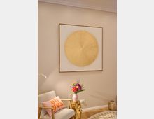 Modern eingerichtetes Wohnzimmer mit hellem Sessel, rosa Kissen mit der Aufschrift „oh yes“ und einer goldenen Beistelltisch-Skulptur. Darüber hängt ein großes, gerahmtes Kunstwerk mit einem strahlenförmigen goldenen Kreis; daneben steht eine Vase mit bunten Blumen.