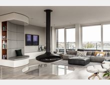 Modernes Wohnzimmer mit hellen Holzböden, großer Fensterfront und einer Hängerkaminanlage aus schwarzem Metall in der Raummitte. Ein graues Ecksofa, ein weißes Sitzmöbel mit Regal und ein runder Glaseinsatz im Boden prägen die Einrichtung.