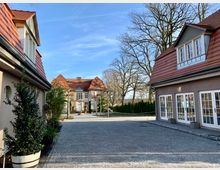 Gepflegter Hofbereich mit gepflastertem Boden, flankiert von zwei Gebäuden mit roten Ziegeldächern und großen Fenstern. Im Hintergrund steht ein größeres Haus im klassischen Stil mit heller Fassade und Zierbäumen, umgeben von hohen Bäumen ohne Laub.