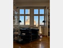Wohnzimmer mit modernem Interieur, bestehend aus einem schwarzen Ledersessel, einem kleinen Tisch mit Büchern und Regalen an den Seitenwänden. Große Fenster mit Ausblick auf ein benachbartes Gebäude mit Turm im Hintergrund, das von Sonnenlicht beleuchtet wird.
