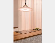 Moderne Küche mit heller Holzarbeitsplatte und glatter, schwarzer Kochfläche. Darüber hängt eine schlichte, runde Pendelleuchte, im Hintergrund sind helle, gefaltete Vorhänge zu sehen.