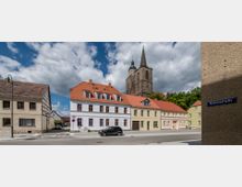 Straßenszene in einem Dorf mit historischen Gebäuden, darunter ein Fachwerkhaus, Häuser mit roten Ziegeldächern und hell gestrichenen Fassaden sowie eine große Kirche mit einem steinernen Turm und Spitzdach im Hintergrund. Der Straßenname "Schulstraße" ist auf einem Schild sichtbar.