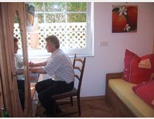 Ein kleiner Wohnraum mit einem Fenster, durch das Tageslicht einfällt und ein weißer Zaun mit grüner Hecke sichtbar ist. Ein Mann sitzt auf einem Stuhl vor einem Spiegel, daneben ein Sofa mit roten Kissen sowie ein Gemälde mit Früchten an der Wand.