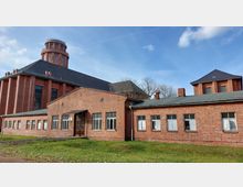 Backsteinbau mit mehreren flach geneigten Dächern und großen Fenstern, daneben ein größeres Gebäude mit einem markanten runden Turm und hohen Fenstern. Der Komplex befindet sich in einer offenen Umgebung mit Rasen im Vordergrund.