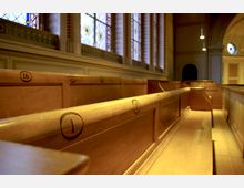 Innenraum einer Kirche mit hölzernen, nummerierten Kirchenbänken im Vordergrund. Im Hintergrund befinden sich bunte, verzierte Glasfenster sowie gewölbte Decken und dezente Beleuchtung.