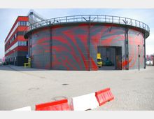Rundes Industriegebäude mit grauer Fassade und roten, abstrakten Mustern sowie einem angrenzenden, mehrstöckigen, rechteckigen Bürogebäude in Rot-Schwarz. Im Vordergrund ist eine gepflasterte Fläche mit rot-weißen Barrieren, und durch ein großes Tor des runden Gebäudes sind zwei Fahrzeuge sichtbar.