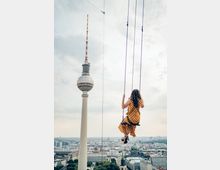 Eine Frau in einem gepunkteten Kleid schaukelt hoch über der Stadt Berlin mit Blick auf den Fernsehturm. Im Hintergrund sind die Skyline Berlins und die grüne Kuppel des Berliner Doms sichtbar.