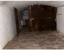 Gewölbekeller mit weiß gestrichenen Ziegelwänden und Backsteinboden, am Ende durch ein großes dunkles Holztor verschlossen. Links steht eine geöffnete Holztür zu einem schmalen Gang mit freiliegenden Leitungen und einem Heizkörper.