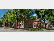 Backsteingebäude in einer parkähnlichen Umgebung mit hohen Bäumen, gepflasterten Wegen und einem klaren blauen Himmel. Die Gebäude haben Satteldächer und weiße Fensterrahmen, die von grünem Laub umrahmt werden.