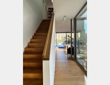 Treppenaufgang aus Holz in einem modernen Innenraum mit Holzboden. Rechts daneben befindet sich eine Glaswand, die ein Wohnzimmer mit Sitzmöbeln und Blick nach draußen in einen sonnigen Garten zeigt.