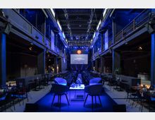 Innenraum einer modernen Veranstaltungsstätte mit industriellem Design, hohen Decken und Stahlstützen. Der Raum ist mit gedeckten Tischen, Stühlen, einer Bühne mit zwei Stühlen im Vordergrund und einer großen beleuchteten Leinwand im Hintergrund ausgestattet.