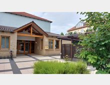 Eingangsbereich eines modernen Gebäudes mit Ziegelsteinfassade, großen Holzfenstern und einem überdachten Eingang mit Holzbalken. Im Vordergrund sind bepflanzte Grünflächen und ein Fahrradständer, rechts steht eine Holzgitterkonstruktion.