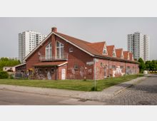 Backsteinbau mit Satteldach und roten Ziegeln in einer Wohngegend, umgeben von Grünflächen und Hochhäusern im Hintergrund; die Fassade ist mit Graffiti besprüht, und ein Straßenschild weist auf den "Rebhuhnweg" hin. Ein gepflasterter Gehweg verläuft entlang der Straße.