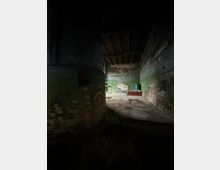 Dunkler, leerer Industrieraum mit großen zylindrischen Mauern, freiliegenden Holzbalken an der Decke und abgenutzten Ziegelwänden mit grünem Anstrich. Im Hintergrund sind Rohrleitungen, eine kleine erhöhte Plattform und ein grüner Behälter auf einem staubigen Betonboden zu sehen.
