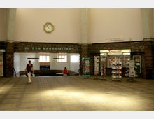 Eingangsbereich eines Bahnhofs mit einem Schriftzug „Zu den Bahnsteigen“ über dem Treppenabgang, einer Wanduhr und einem Zeitungs- und Bücherladen auf der rechten Seite. Die Wände sind teils mit dunkelbraunen Fliesen verkleidet und der Raum ist von Tageslicht erhellt.