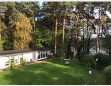 Einstöckiges Gebäude mit großen Glasfenstern, umgeben von einem gepflegten grünen Rasen und hohen Nadelbäumen. Im Hintergrund sind ein See und dichte Vegetation zu sehen, mit vereinzelten Fahnen im Garten.