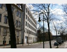Straßenszene mit einem mehrstöckigen, hellen Gebäude auf der linken Seite, Bäumen ohne Laub entlang des Gehwegs und einer leeren Straße mit parkenden Autos. Im Hintergrund ist eine Brücke und teils bewölkter Himmel zu sehen.