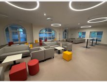 Ein moderner Aufenthaltsraum mit grauen Sofas, weißen Tischen und bunten Hockern in Rot und Gelb. Große Fenster mit Rundbögen lassen Tageslicht ein, und runde, leuchtende Deckenleuchten hängen von der weißen Decke.