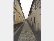 Eine schmale, gepflasterte Gasse mit beidseitigen Wohnhäusern. Auf der linken Seite sind verputzte Fassaden, rechts ein Fachwerkgebäude; am Ende der Straße ist ein Backsteingebäude zu sehen.