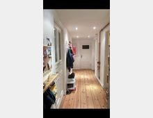 Ein schmaler Flur in einer Wohnung mit Holzdielenboden und weißen Wänden. Links an der Wand befinden sich eine Tür, ein Regal mit Haken und Ablage sowie eine Pinnwand, während rechts ein offenes Regal sichtbar ist; im Hintergrund eine weiße Tür mit Glasfenster.