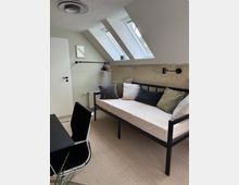 Helles Arbeits- oder Gästezimmer mit schräger Decke und drei Dachfenstern. Ein Metallbett mit Kissen steht an der Wand, daneben ein kleiner schwarzer Schreibtisch mit Bürostuhl; die Wand ist teils verputzt und teils gestrichen.