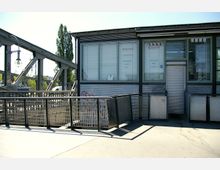 Metallbauwerk mit Geländern und einem kleinen Gebäude. Das Gebäude hat große Fenster mit blickdichten Jalousien, französische Elemente in der Beschriftung und eine geschlossene Rollladentür. Im Hintergrund sind ein Stahlträger mit Straßenlampe sowie Bäume sichtbar.