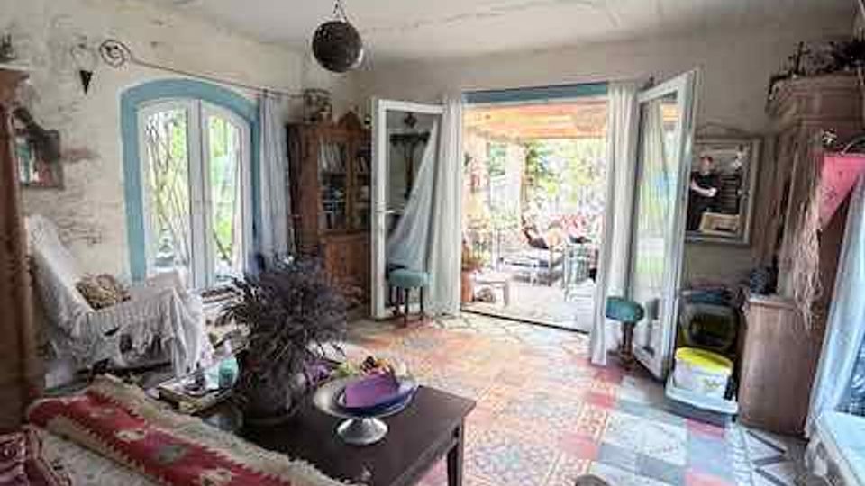 Rustikales Wohnzimmer mit abgenutztem Putz an der Decke, gemustertem Fliesenboden und verschiedenen Holzmöbeln sowie Sitzgelegenheiten. Große Flügeltüren und Fenster lassen Tageslicht herein und führen auf eine überdachte Terrasse mit Blick ins Grüne.