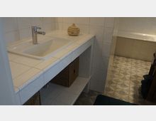 Badezimmer mit einem weißen Waschbecken auf einer gefliesten Ablage, darunter offene Regalfächer mit geflochtenen Körben. Im Hintergrund ist eine geflieste Badewanne und gemusterter Bodenbelag zu sehen.