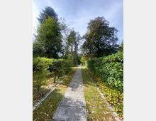 Ein gerader Weg aus rechteckigen Steinplatten führt durch einen Garten, gesäumt von grünenden Büschen und Hecken. Im Hintergrund erheben sich hohe Bäume, die eine parkähnliche Atmosphäre schaffen, unter einem leicht bewölkten Himmel.