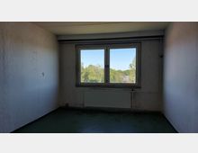 Leerer Raum mit weißen, leicht abgenutzten Wänden und einem Fenster mit Blick auf Bäume und blauen Himmel. Unter dem Fenster befindet sich ein Heizkörper, der Boden ist dunkel und zeigt Gebrauchsspuren.