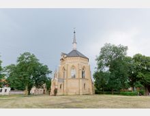 Kirche im neugotischen Stil mit hellbrauner Fassade, spitzbogigen Fenstern und einem hohen Turm mit Spitzhelm, umgeben von Rasen und Bäumen; im Hintergrund kleine Gebäude und ein bewölkter Himmel.