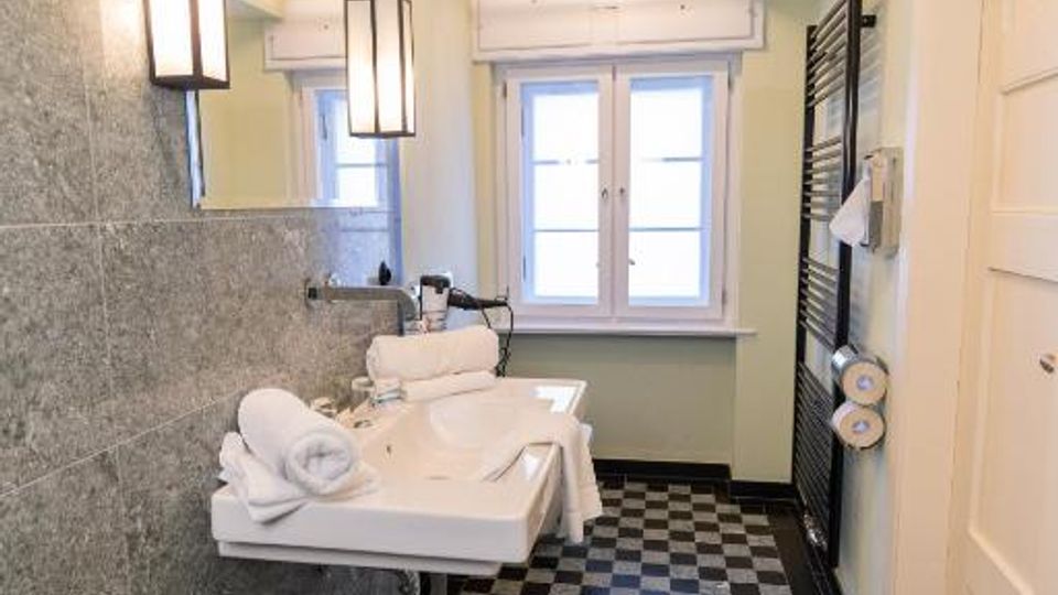 Schmales Badezimmer mit schwarz-weiß kariertem Fliesenboden, grauer Wandverkleidung und langem Spiegel über einem wandmontierten Waschbecken. Am Ende des Raums steht ein Doppelfenster, rechts sind ein schwarzer Handtuchheizkörper und eine weiße Tür zu sehen.