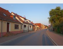 Straße in einem Wohngebiet mit Reihen von Einfamilienhäusern im traditionellen Stil, gelblich-beigen Fassaden und roten Ziegeldächern. Die Straße ist asphaltiert, mit Gehwegen und vereinzelt Laternen auf beiden Seiten.