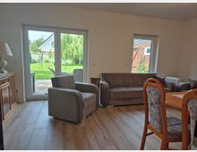 Helles Wohnzimmer mit Holzfußboden, grauem Sessel und Sofa sowie einem angrenzenden Essbereich. Große Terrassentür und ein Seitenfenster bieten Blick in den Garten mit Rasen und Terrasse.