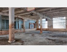 Innenansicht einer verlassenen Fabrikhalle mit freiliegender Betonstruktur, rostigen Säulen und großen, rechteckigen Fenstern. Die Wände zeigen Abnutzungsspuren, teilweise mit abblätternder Farbe, und der Boden ist aus Beton.